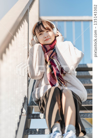 年輕女子坐在樓梯上,戴著耳機聽音樂。 年輕女子坐在樓梯上,戴著耳機聽音樂。 135143302