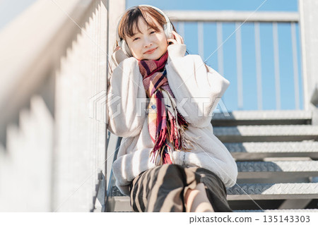 年輕女子坐在樓梯上,戴著耳機聽音樂。 年輕女子坐在樓梯上,戴著耳機聽音樂。 135143303