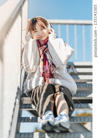 年輕女子坐在樓梯上,戴著耳機聽音樂。 年輕女子坐在樓梯上,戴著耳機聽音樂。 135143305