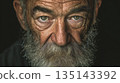 Intense portrait of an elderly man with a weathered face, grey beard, and piercing green eyes set against a dark background. His gaze reflects experience & wisdom. 135143392