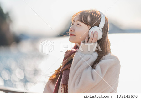 一位年輕女子站在海邊，戴著耳機聽音樂。 135143676