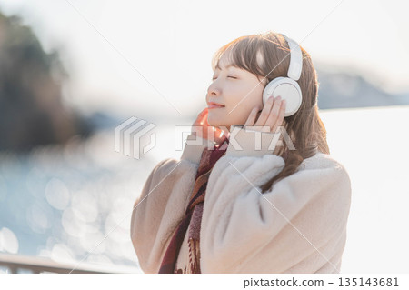 Young woman standing by the seaside listening to music with headphones Young woman standing by the seaside listening to music with headphones 135143681
