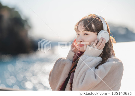 一位年輕女子站在海邊，戴著耳機聽音樂。 135143685