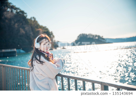 一位年輕女子站在海邊，戴著耳機聽音樂。 135143694
