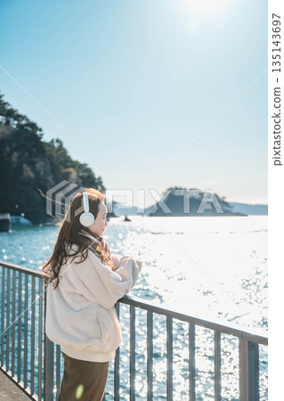 一位年輕女子站在海邊，戴著耳機聽音樂。 135143697