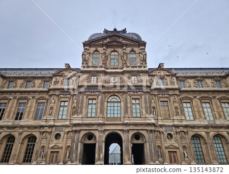 Paris, France - December 10, 2025: The ornate Renaissance facade and the iconic glass Pyramid of the Louvre Museum with multiple tourists scattered across the vast cobblestone courtyard 135143872