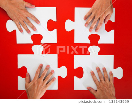 Four hands positioning white puzzle pieces on a red background 135143969