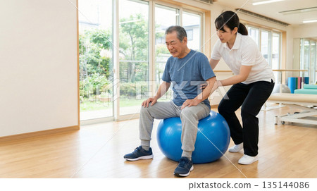 An elderly man works hard at rehabilitation using a balance ball while a physical therapist watches over him. A training scene at a nursing home. An elderly man works hard at rehabilitation using a balance ball while a physical therapist watches over him. A training scene at a nursing home. 135144086