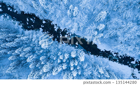 Aerial top down view of a snow covered winter forest with a frozen stream cutting through frosted pine trees. Minimal, serene winter landscape captured by drone in cold blue tones. 135144280