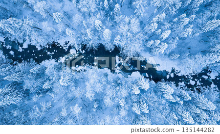 Aerial top down view of a snow covered winter forest with a frozen stream cutting through frosted pine trees. Minimal, serene winter landscape captured by drone in cold blue tones. Aerial top down view of a snow covered winter forest with a frozen stream cutting through frosted pine trees. Minimal, serene winter landscape captured by drone in cold blue tones. 135144282