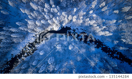 Aerial top down view of a snow covered winter forest with a frozen stream cutting through frosted pine trees. Minimal, serene winter landscape captured by drone in cold blue tones. Aerial top down view of a snow covered winter forest with a frozen stream cutting through frosted pine trees. Minimal, serene winter landscape captured by drone in cold blue tones. 135144284
