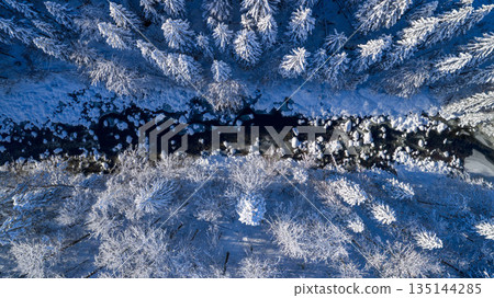 Aerial top down view of a snow covered winter forest with a frozen stream cutting through frosted pine trees. Minimal, serene winter landscape captured by drone in cold blue tones. Aerial top down view of a snow covered winter forest with a frozen stream cutting through frosted pine trees. Minimal, serene winter landscape captured by drone in cold blue tones. 135144285