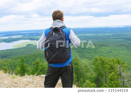 A man in a blue tank top and gray turtleneck stands on a mountain top with a backpack. A man in a blue tank top and gray turtleneck stands on a mountain top with a backpack. 135144559