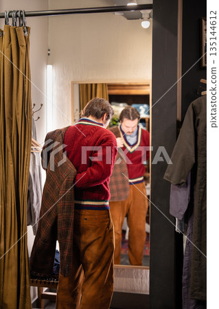 Hipster man customer fits on wool second hand jacket in front of mirror in fancy vintage shop. 135144612