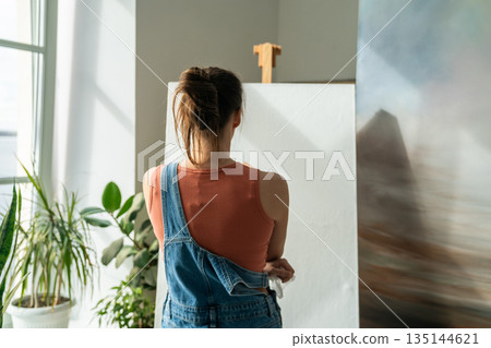 Serious female artist painting masterpiece at home loft studio, thinking and looking at empty canvas Serious female artist painting masterpiece at home loft studio, thinking and looking at empty canvas 135144621