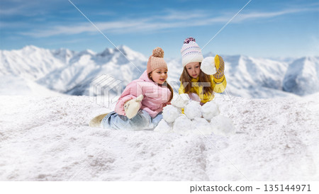 Playful artwork of two girls building snow structure together in mountains. Playful artwork of two girls building snow structure together in mountains. 135144971