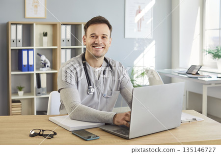 Portrait of a young happy male doctor or medical nurse in uniform sitting at the desk in his office Portrait of a young happy male doctor or medical nurse in uniform sitting at the desk in his office 135146727