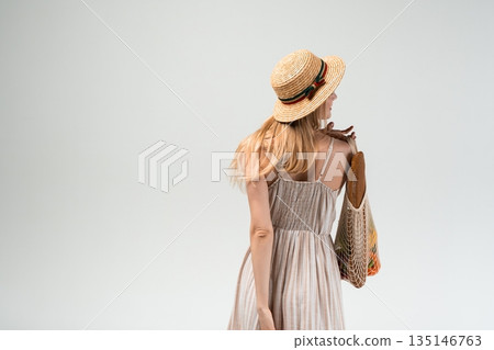 Young woman seen from behind wearing straw hat and summer dress carrying reusable mesh bag with vegetables, eco lifestyle concept Young woman seen from behind wearing straw hat and summer dress carrying reusable mesh bag with vegetables, eco lifestyle concept 135146763