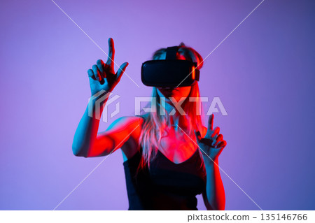 Young woman playing VR gaming with headset, pointing and interacting in virtual reality, neon red blue studio lighting 135146766