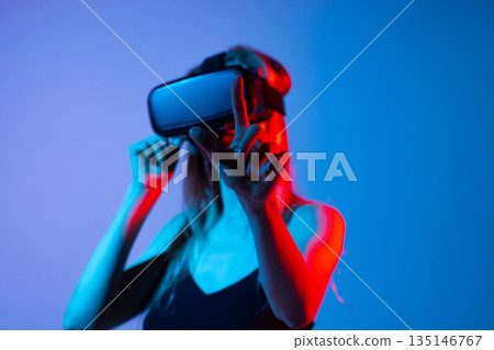 Female gamer using virtual reality headset and touching virtual interface in neon blue and red light, future technology concept 135146767
