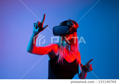 Woman touching virtual interface while wearing VR headset in neon light, immersive digital experience and futuristic technology interaction concept Woman touching virtual interface while wearing VR headset in neon light, immersive digital experience and futuristic technology interaction concept 135146769
