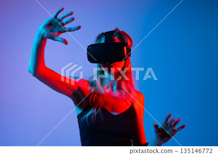 Female experiencing virtual reality using VR headset and expressive hand movements, neon lighting and futuristic immersive digital technology concept Female experiencing virtual reality using VR headset and expressive hand movements, neon lighting and futuristic immersive digital technology concept 135146772
