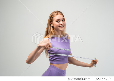 Smiling young woman in purple sportswear holding measuring tape, body measurement and weight loss concept on gray studio background 135146792