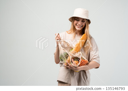 Young woman in bucket hat carefully holding reusable mesh grocery bag with vegetables and bread, sustainable lifestyle concept 135146793