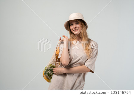 Young woman in bucket hat carrying reusable mesh shopping bag over shoulder, sustainable lifestyle and eco shopping concept Young woman in bucket hat carrying reusable mesh shopping bag over shoulder, sustainable lifestyle and eco shopping concept 135146794