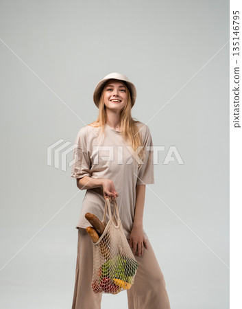 Smiling young woman in bucket hat holding reusable mesh grocery bag with vegetables and bread, zero waste shopping concept 135146795