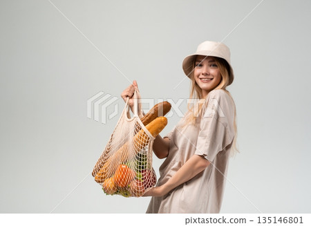 Cheerful young woman with reusable grocery bag full of bread and vegetables, sustainable shopping and healthy lifestyle concept 135146801