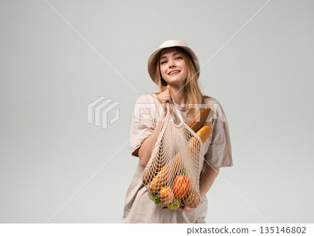 Smiling young woman in bucket hat holding reusable mesh grocery bag with bread and fresh produce, eco lifestyle portrait 135146802