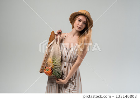 Stylish young woman holding eco friendly grocery bag with fresh produce, straw hat and summer dress isolated on gray background Stylish young woman holding eco friendly grocery bag with fresh produce, straw hat and summer dress isolated on gray background 135146808