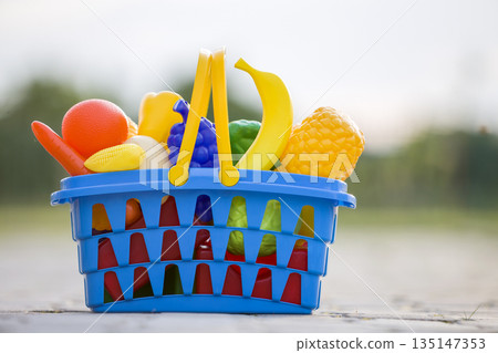 Bright plastic colorful basket with toy fruits and vegetables outdoors on sunny summer day. 135147353