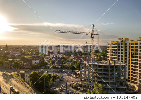 Apartment or office tall building under construction. Brick walls, glass windows, scaffolding and concrete support pillars. Tower crane on bright blue sky copy space background. 135147402