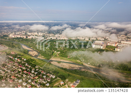 Aerial view of white clouds above a town or village with rows of buildings and curvy streets between green fields in summer. Countryside landscape from above. 135147428