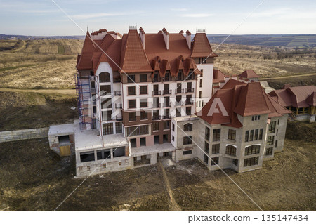 Aerial view of unfinished multistoried residence, hotel or cottage building with stucco wall, cast iron balcony railings, steep shingle roof and shiny windows on rural landscape background. Aerial view of unfinished multistoried residence, hotel or cottage building with stucco wall, cast iron balcony railings, steep shingle roof and shiny windows on rural landscape background. 135147434