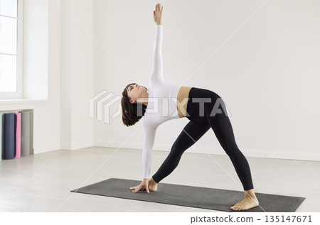 Woman performing triangle yoga pose on mat, reaching upward in serene studio 135147671