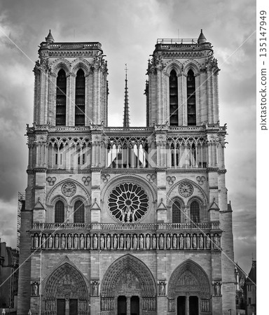 The Notre-Dame Cathedral facade in Paris, France. Black and white view of the intricate portals, the Gallery of Kings, the iconic rose window and two massive bell towers against a dramatic sky 135147949