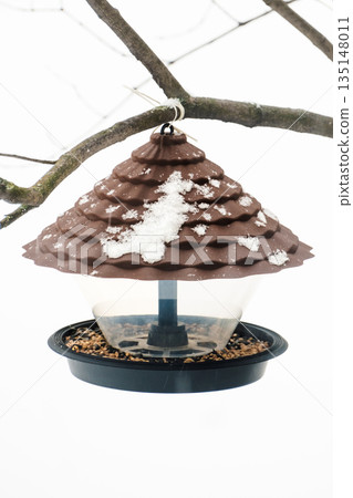 A plastic bird feeder with seeds and bird food hangs from a tree. Caring for birds in winter 135148011