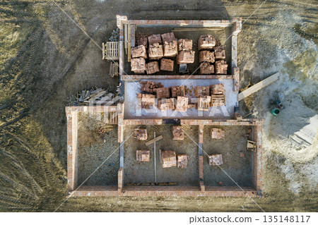 Aerial view of building site. Trenches dug in ground and filled with cement as foundation for future house, brick basement floor and stacks of brick for construction. Aerial view of building site. Trenches dug in ground and filled with cement as foundation for future house, brick basement floor and stacks of brick for construction. 135148117