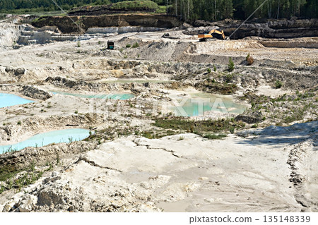 Digging excavator on a quarry with kaolin clay, light sand and blue water, beautiful landscape, industrial excavation of useful minerals Digging excavator on a quarry with kaolin clay, light sand and blue water, beautiful landscape, industrial excavation of useful minerals 135148339