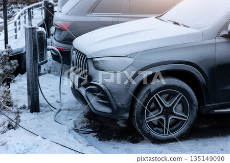 Electric vehicle charging outdoors winter conditions with snow frost covering car and charger highlighting cold weather impact battery performance, range efficiency everyday electric mobility usage 135149090
