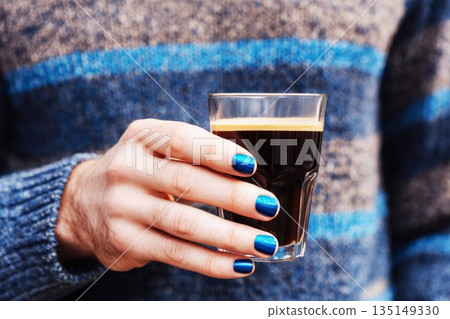 A man sits in a comfortable position, holding a blue coffee cup. His nails are recently manicured in a dark color. He wears a knitted sweater and jeans, creating a casual look 135149330