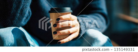 A man sits in a comfortable position, holding a blue coffee cup. His nails are recently manicured in a dark color. He wears a knitted sweater and jeans, creating a casual look, banner 135149331