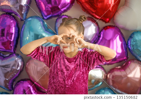 The girl looks through her fingers, folded in the shape of a heart. Congratulations and greetings on Valentine's Day. The girl looks through her fingers, folded in the shape of a heart. Congratulations and greetings on Valentine's Day. 135150682