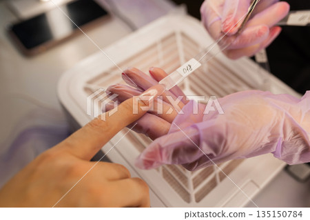 Close up of professional manicure color selection using nail swatch. Nail technician wearing gloves helping female client choose nude gel polish shade in beauty salon. Close up of professional manicure color selection using nail swatch. Nail technician wearing gloves helping female client choose nude gel polish shade in beauty salon. 135150784