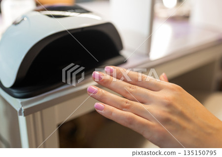 Close up of female hand curing gel nail polish under UV LED lamp in beauty salon. Professional manicure process, nail drying, cosmetic treatment, hygiene and nail care concept. 135150795
