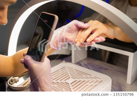 Nail technician photographing finished manicure on female hands using smartphone under ring lamp in beauty salon. Professional nail care, content creation, social media and manicure service concept. 135150796