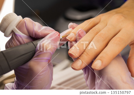 Close up of professional manicure procedure using electric nail drill. Nail technician wearing gloves treating female nails in beauty salon. Nail care, hygiene, cosmetic treatment concept. Close up of professional manicure procedure using electric nail drill. Nail technician wearing gloves treating female nails in beauty salon. Nail care, hygiene, cosmetic treatment concept. 135150797
